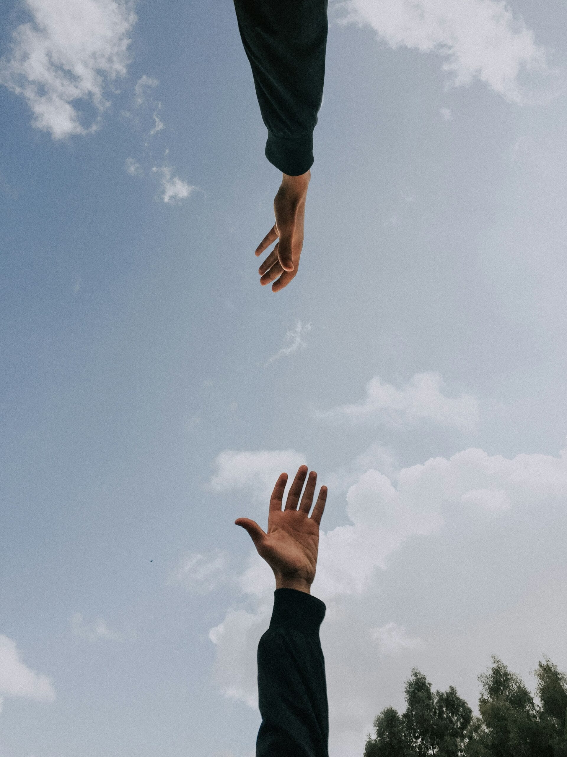 Two hands reaching toward each other against a blue sky with some clouds, symbolizing connection and support—echoing the essence of occupational therapy services. Trees are visible at the bottom edge of the image.